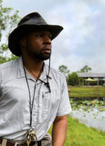 A man in a wide-brimmed hat and gray shirt stands outdoors near a pond, with trees and a building visible in the background.
