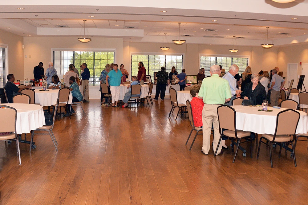 People stand and converse around round tables in a large, well-lit event hall with wooden floors and windows in the background, reflecting the Central Florida Animal Reserve transparency and open community spirit.