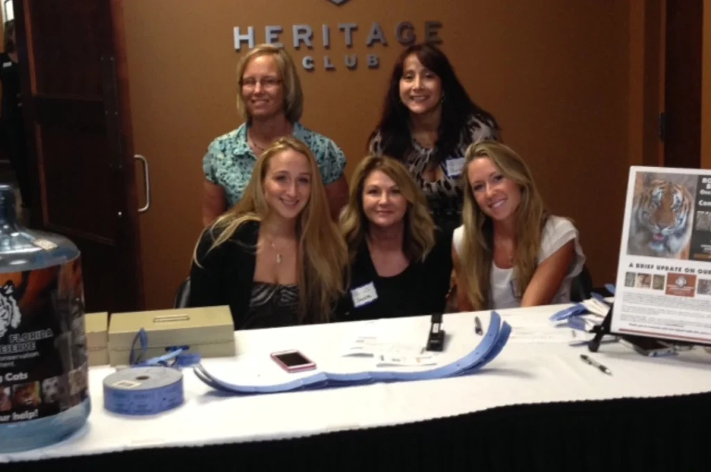 Five women are seated and standing behind a table with raffle tickets, a donation jar, and informational materials about the CFAR partner program at an event in the Heritage Club.