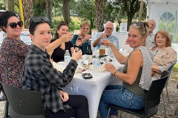 Eight people sit around a white table outdoors, raising glasses and smiling at the camera, with trees and a tent in the background.