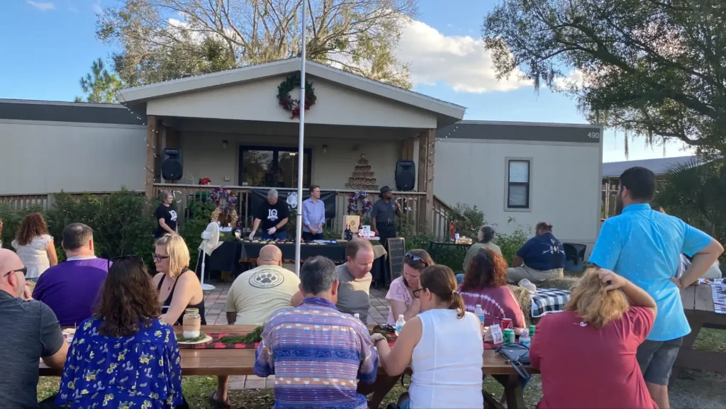 People sit at picnic tables eating, while a group of people stand on a porch in front of a decorated building during an outdoor gathering under a partly cloudy sky.