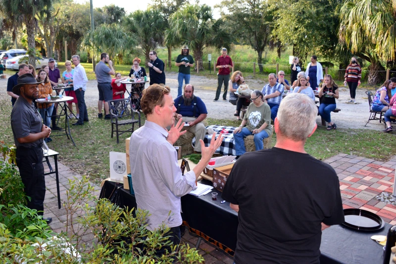 A group of people gather outdoors in Orlando around a speaker who is addressing the audience; some are seated while others stand or listen nearby, enjoying one of the many outdoor activities Orlando has to offer.