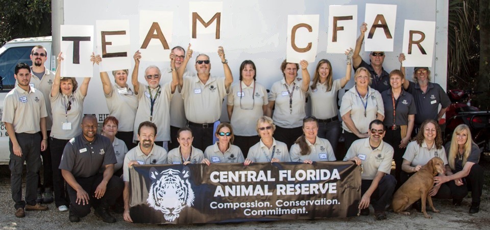 A group of people in matching shirts hold up “TEAM CFAR” signs and a banner that reads “Central Florida Animal Reserve: Compassion. Conservation. Commitment.” A dog sits at the front, representing the spirit of the CFAR partner program.