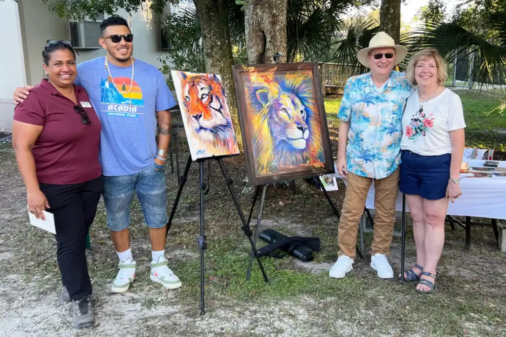 Four people stand outdoors beside two animal paintings on easels, with trees and a building in the background. A table with art supplies, part of the CFAR partner program event, is visible to the right.