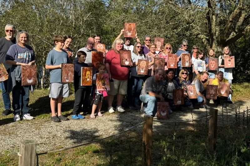 A group of people outdoors holding up wooden boards with painted animal faces, standing on a gravel path surrounded by trees—perfect for those looking to get involved with Central Florida Animal Reserve.