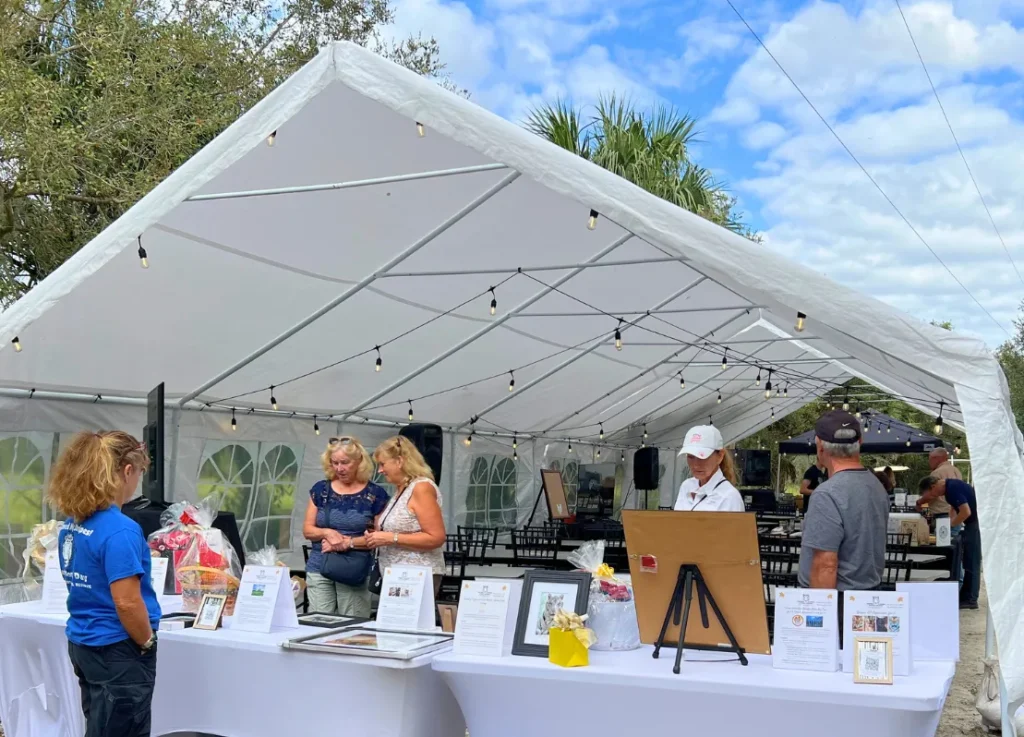People stand under a large white event tent with tables displaying gift baskets and informational materials on a sunny day.
