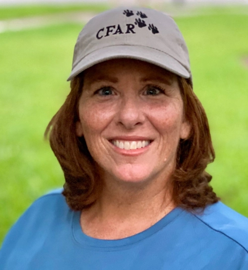 Woman with red hair wears a gray "CFAR" cap and blue shirt, smiling outdoors with green grass behind her—perfect for the Central Florida Animal Reserve media kit.