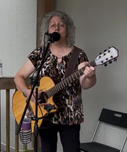 A woman with curly gray hair plays an acoustic guitar and sings into a microphone while standing indoors near a black folding chair.