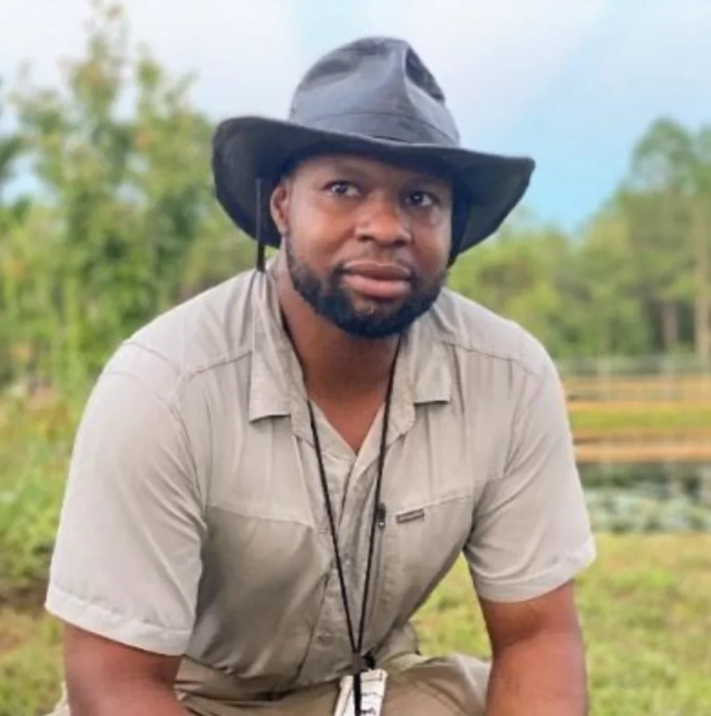 A man wearing a black wide-brim hat and beige shirt, reflecting Central Florida Animal Reserve leadership, crouches outdoors in a grassy area with trees in the background.