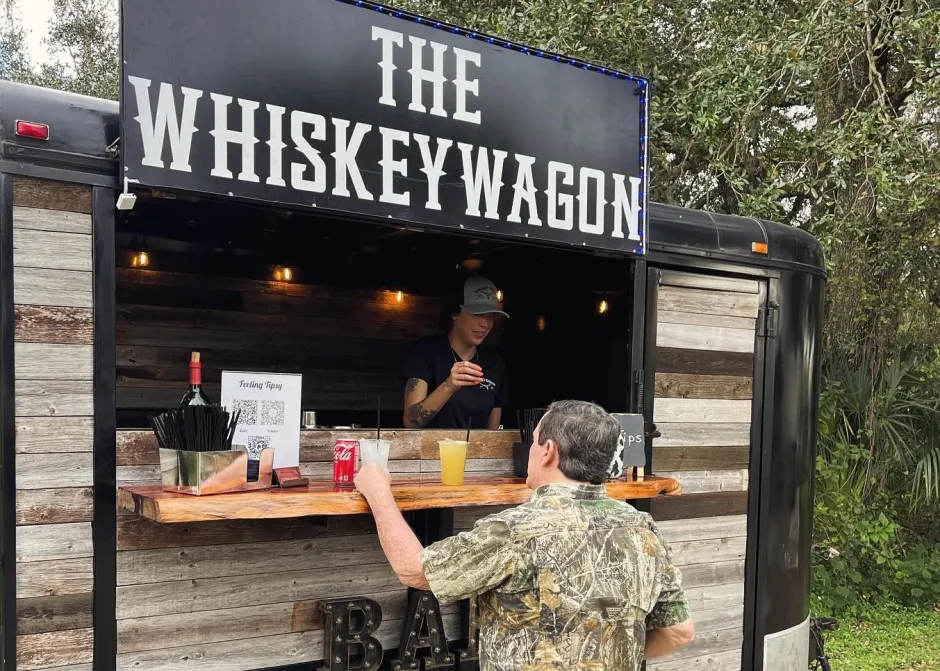 A man orders drinks from a bartender at a rustic outdoor bar trailer labeled "The Whiskey Wagon.