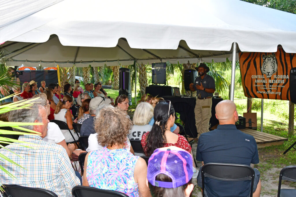 A speaker addresses a seated audience under a tent at the Central Florida Animal Reserve; a banner with a tiger logo is displayed behind the speaker.