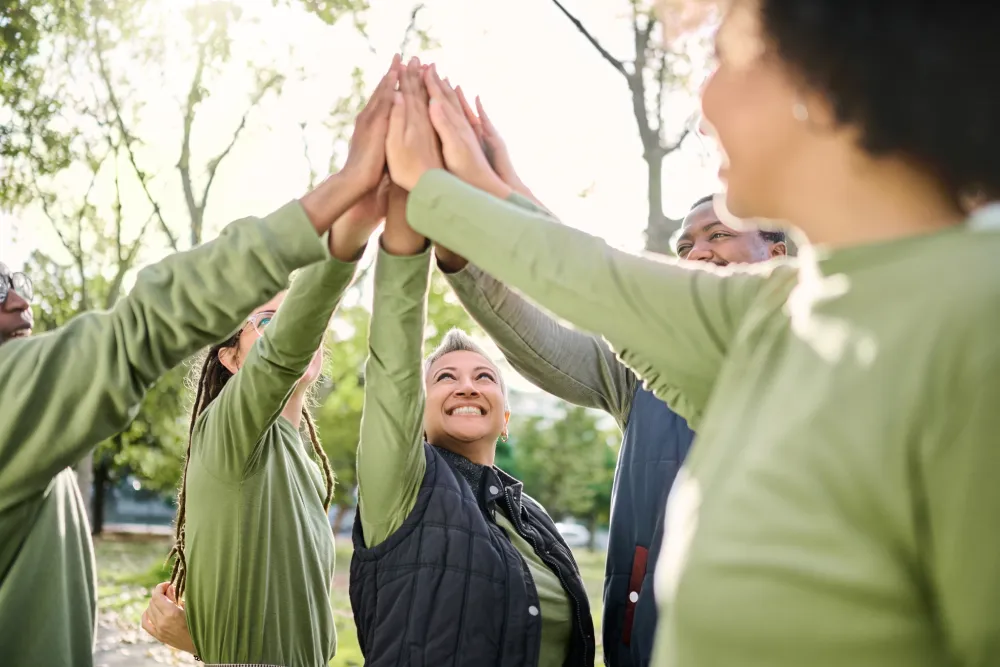 A group of people outdoors, wearing green tops, standing in a circle and joining hands for a group high five.