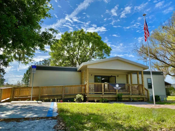 Single-story building with a wooden ramp and deck, an American flag on a pole in front, and trees in the background under a partly cloudy sky—site of the CFAR 3 V’s Campaign.