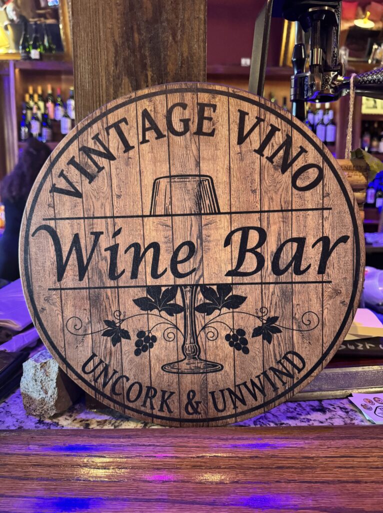 Circular wooden sign for "Vintage Vino Wine Bar" with grapes and wine glass imagery.
