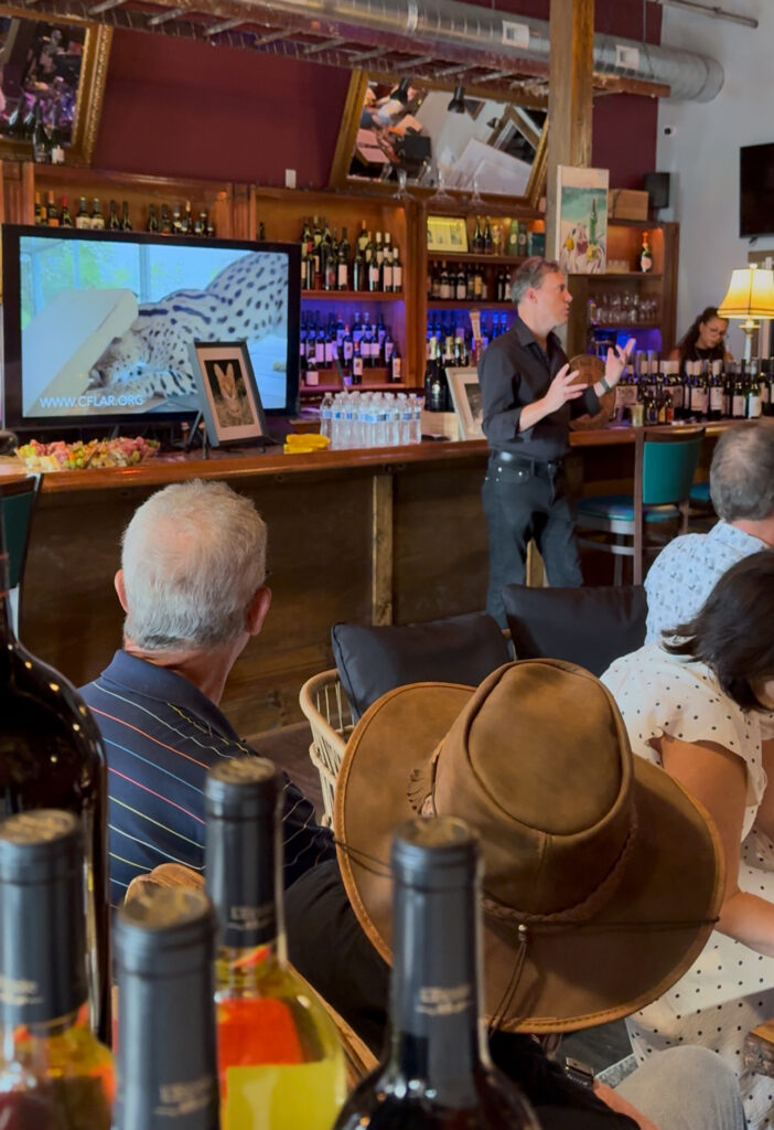 A group of people are gathered in a bar area. A man in a black shirt is presenting, with a screen showing a serval cat behind him. Bottles of wine are in the foreground.